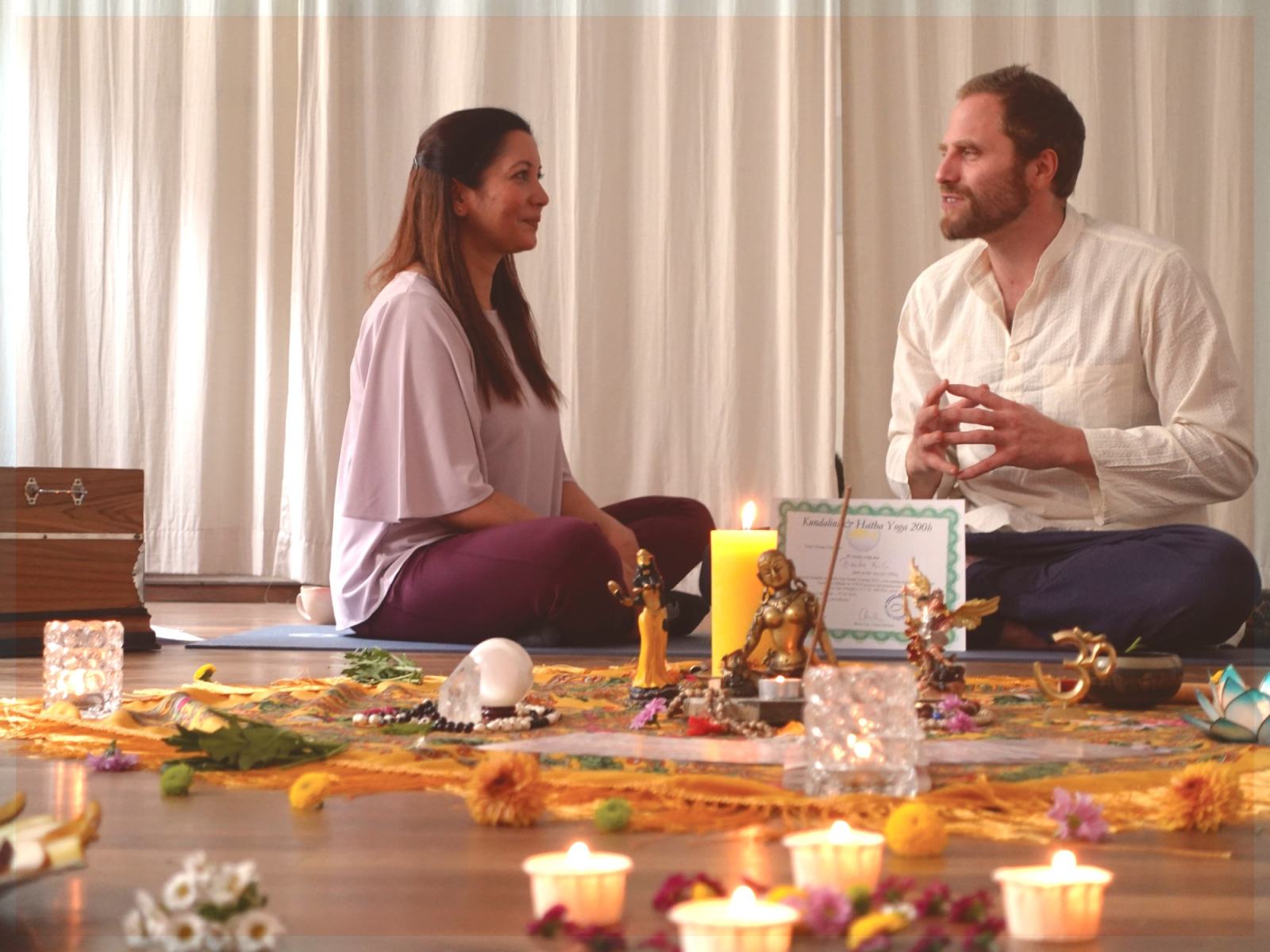 Zwei Yogalehrende sitzen im Gespräch vor einem Altar mit Kerzen und Blumen